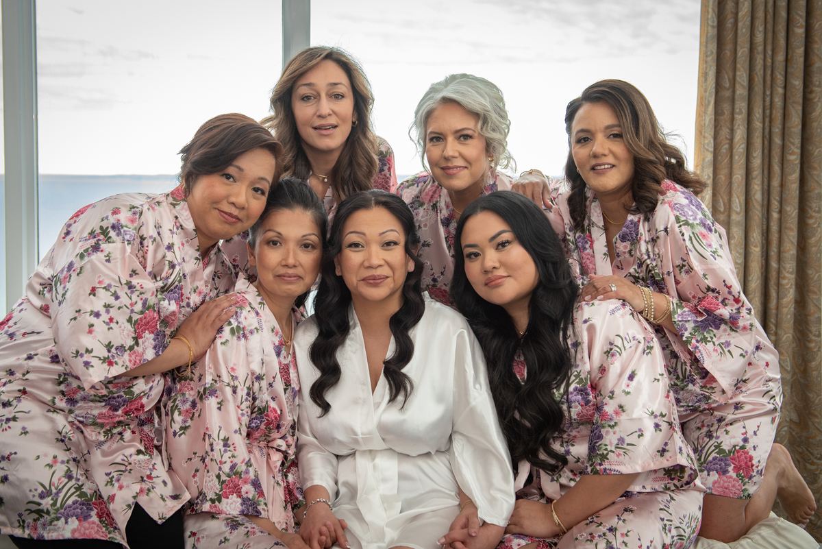 Seven women in pink floral robes and one in a white satin robe gather joyfully near a window with a coastal view.