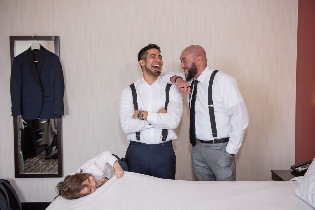 Three men in formal wear laugh together near a bed where a child smiles, with a tuxedo jacket hanging nearby.