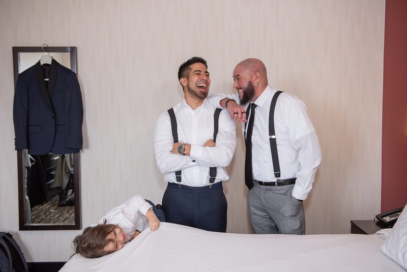 Three men in formal wear laugh together near a bed where a child smiles, with a tuxedo jacket hanging nearby.