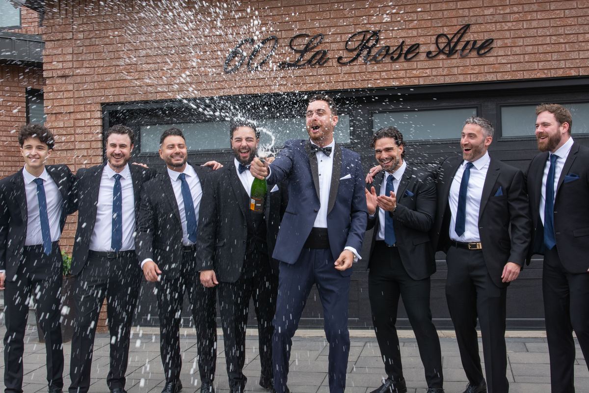 Groomsmen in suits celebrate outside a building, one popping champagne as they laugh and enjoy the moment.