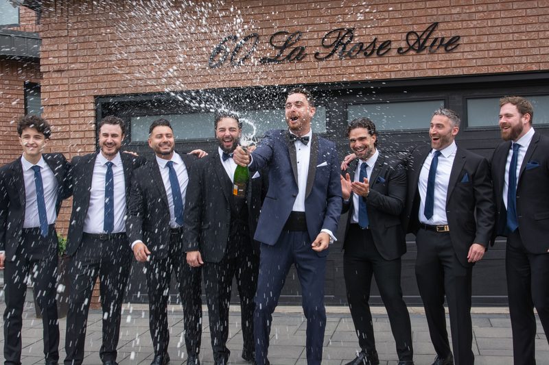 Groomsmen in suits celebrate outside a building, one popping champagne as they laugh and enjoy the moment.
