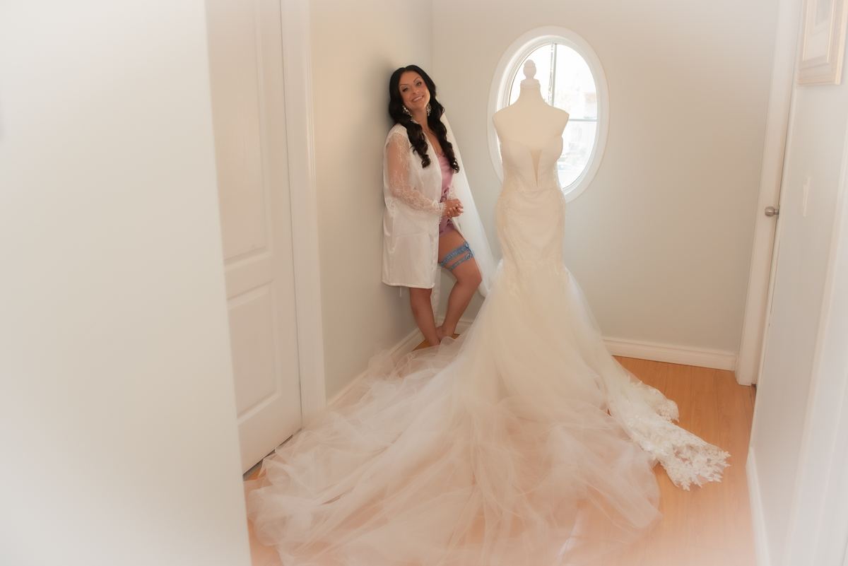 A bride-to-be smiles warmly while leaning against a wall beside a mannequin displaying her long, flowing wedding gown.