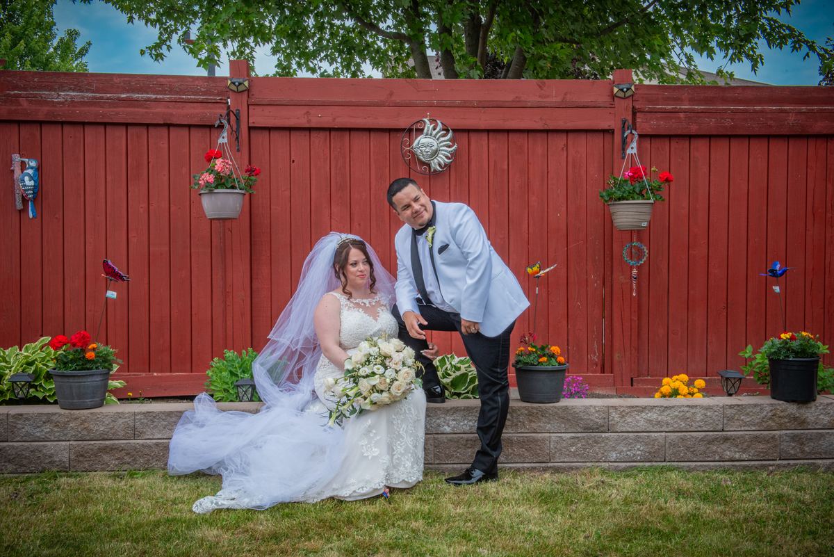 A bride and groom pose playfully in a backyard with a red wooden fence and hanging planters behind them.