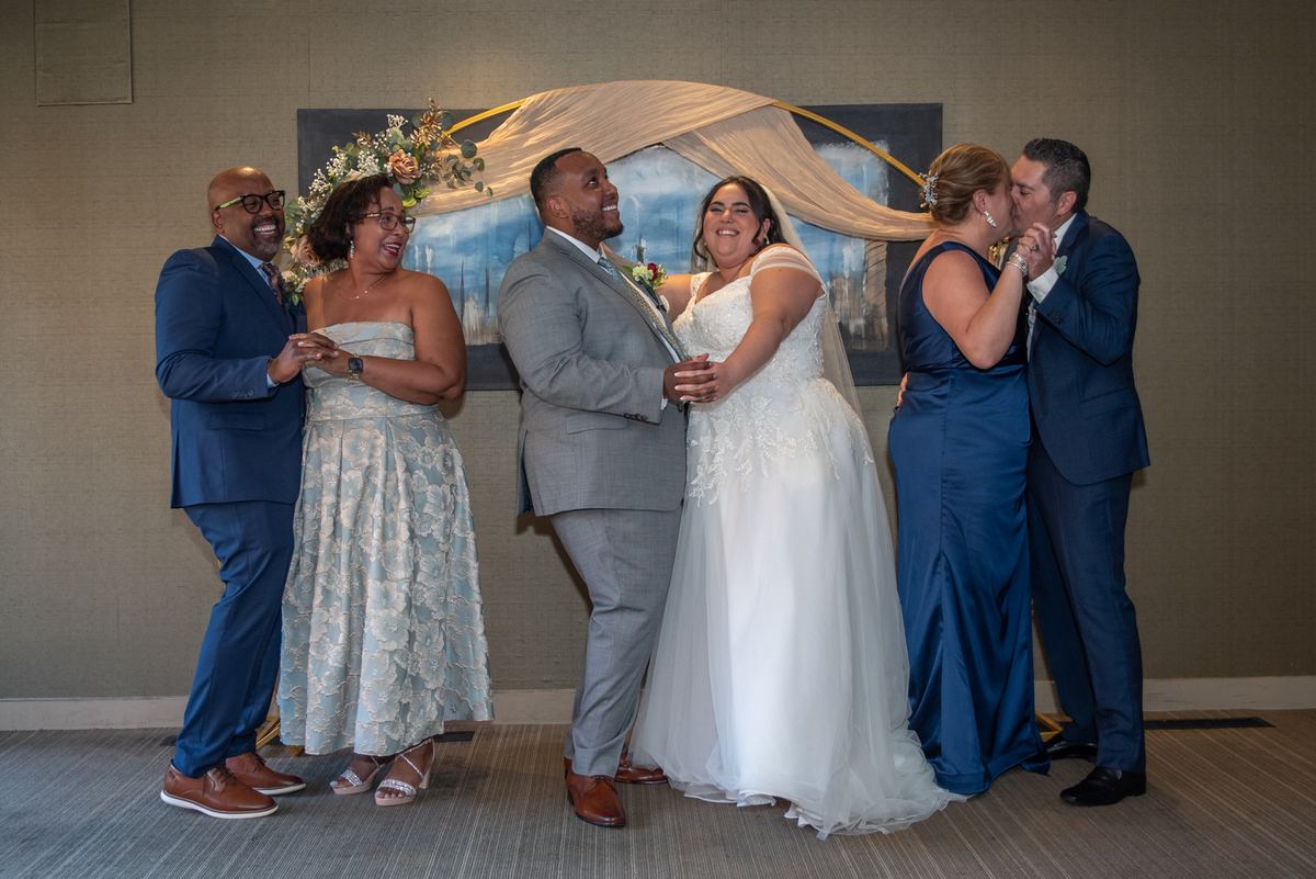 Bride and groom embrace while wedding party shares affectionate kisses under a floral-draped arch, indoors.