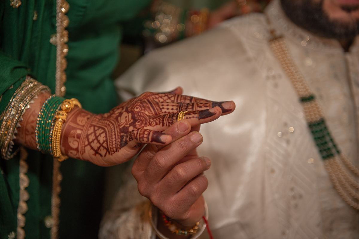 A bride's henna-decorated hand with gold bangles held by the groom in warm lighting during a wedding ritual.