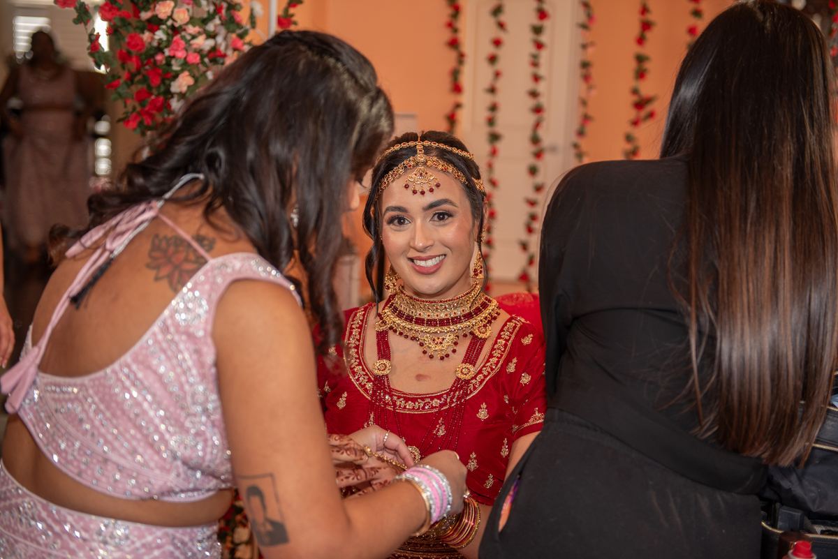 A bride in a red lehenga and gold jewelry smiles as one woman applies henna to her hands while another observes.