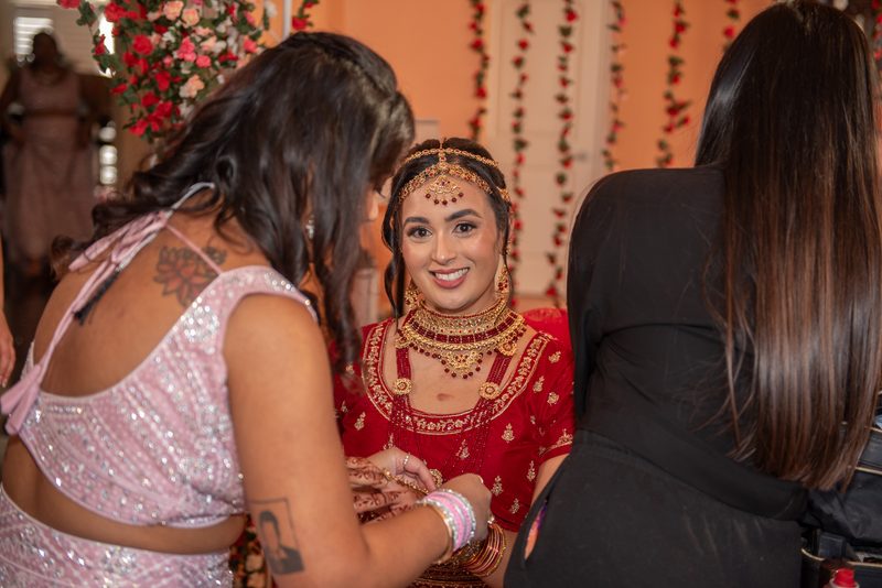 A bride in a red lehenga and gold jewelry smiles as one woman applies henna to her hands while another observes.