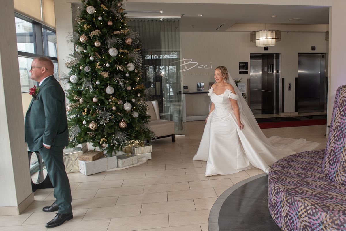 A bride in a white gown approaches a smiling groom near a decorated Christmas tree in a hotel lobby.