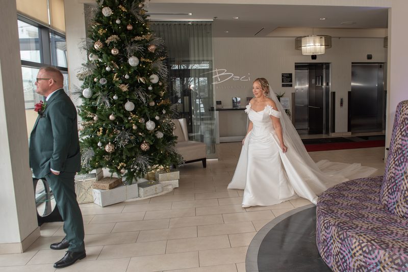 A bride in a white gown approaches a smiling groom near a decorated Christmas tree in a hotel lobby.