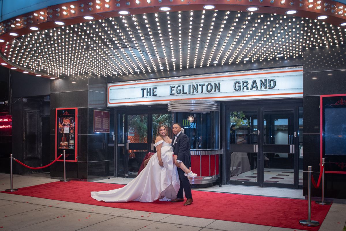 A newlywed couple in formal attire pose joyfully outside an illuminated entrance, the groom lifting the bride's hand.