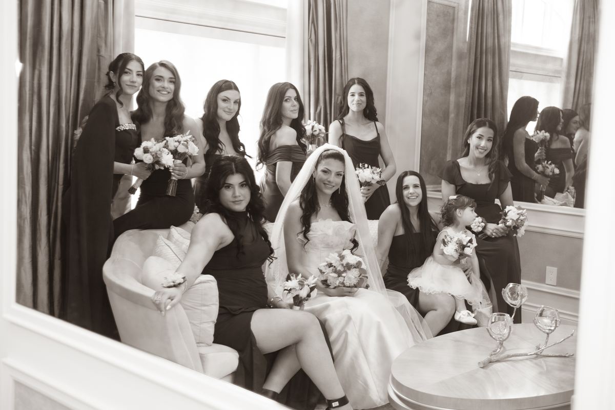 Bride and bridesmaids smile with bouquets in an elegant indoor setting, with a young flower girl beside the bride.