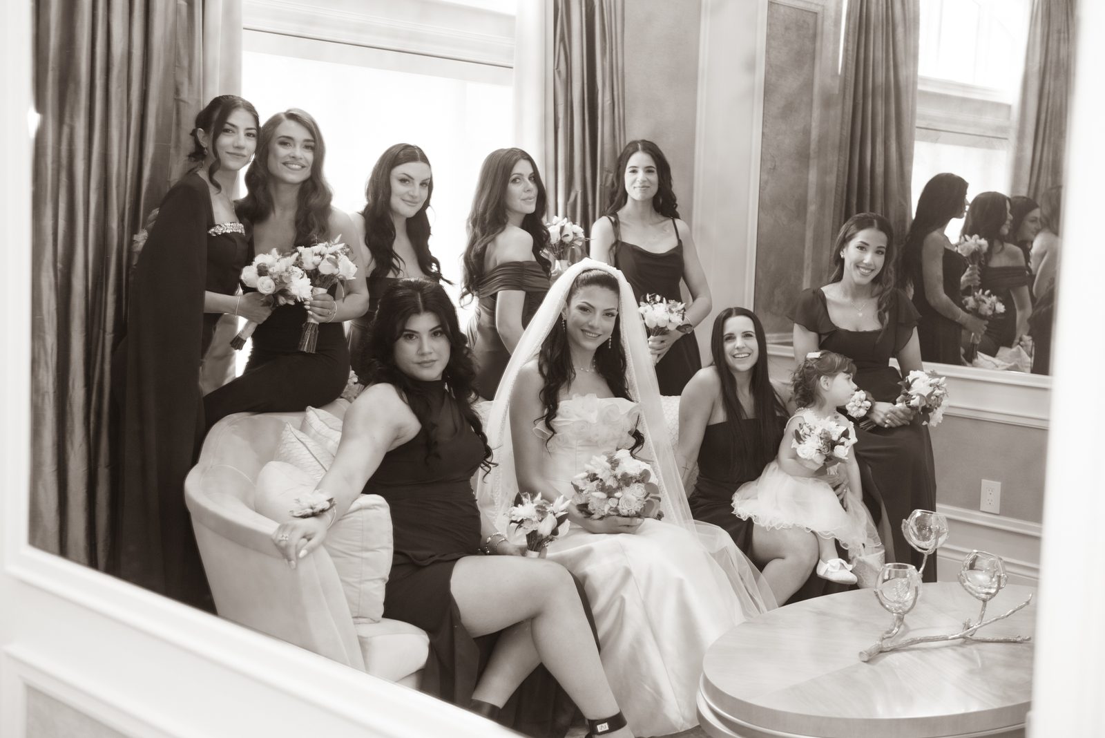 Bride and bridesmaids smile with bouquets in an elegant indoor setting, with a young flower girl beside the bride.