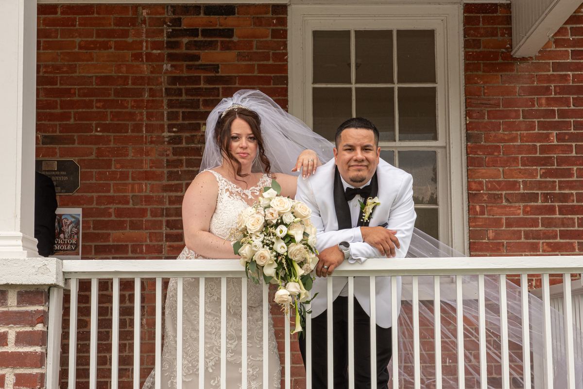 A bride and groom stand on a white-railed porch, the bride holding roses as they look toward the camera.