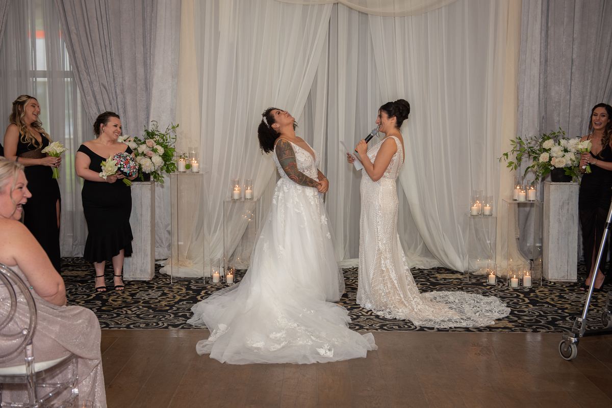 Two brides exchange words at a candlelit ceremony, one laughing, with guests and curtains in the background.