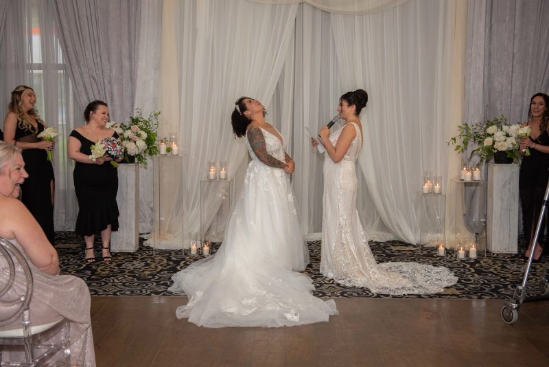 Two brides exchange words at a candlelit ceremony, one laughing, with guests and curtains in the background.