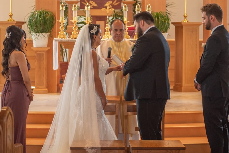 A bride and groom exchange vows at an altar with wooden pews, golden candelabras, and potted plants in the background.