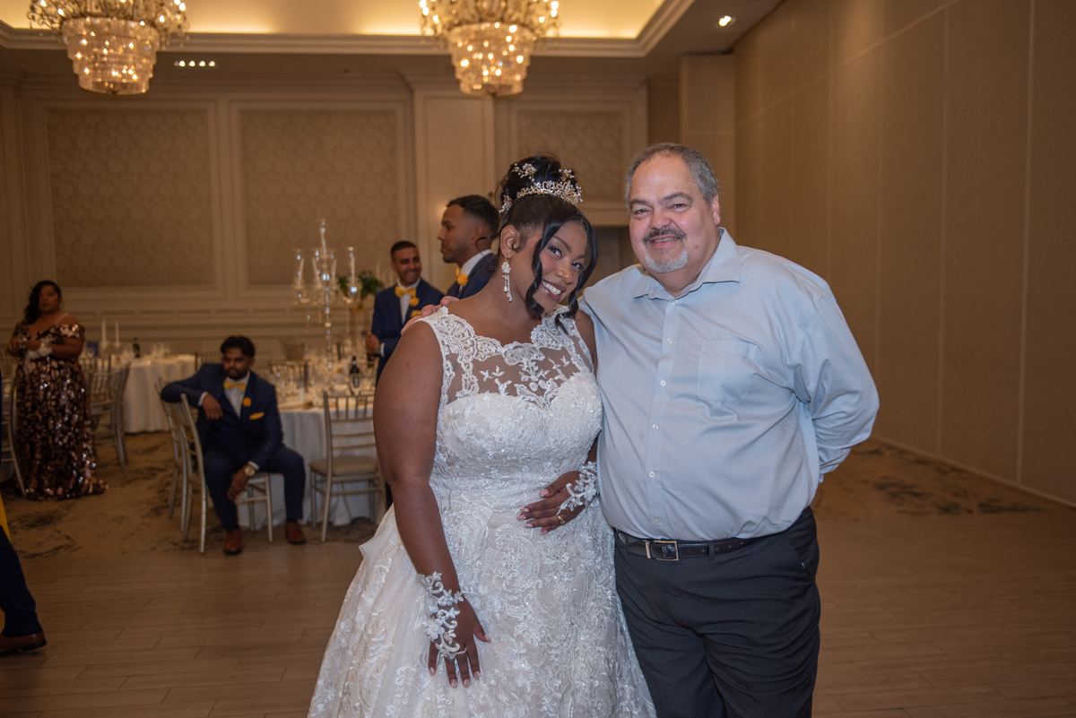 A smiling bride in a white lace gown hugs her father beside an elegantly set table at a wedding reception.