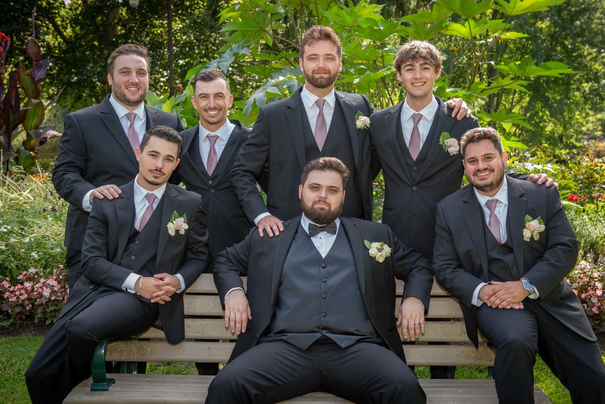 A groom and his groomsmen pose in a garden around a wooden bench, bathed in soft daylight filtering through trees.