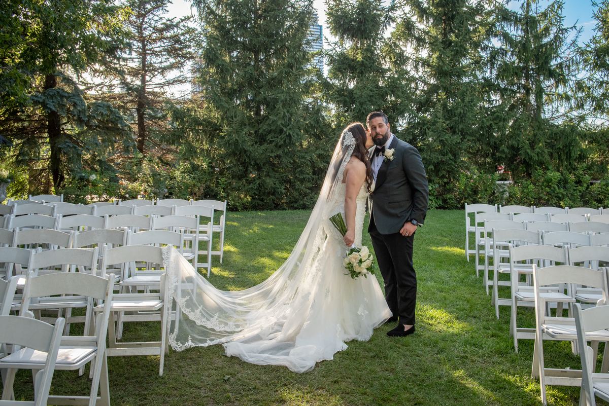 A bride in a white gown and veil leans to kiss her groom's cheek as he holds a bouquet of white flowers.