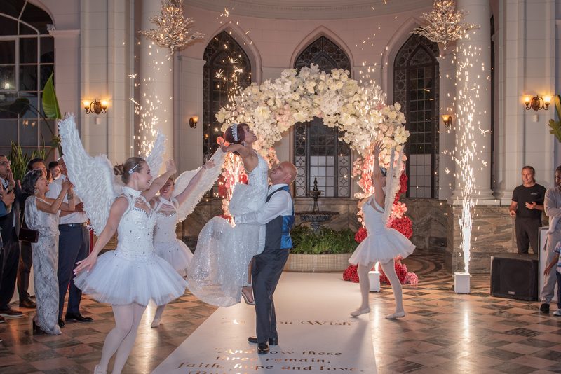 The bride is joyfully lifted by her groom under a floral arch, surrounded by dancers and fireworks in a grand hall.
