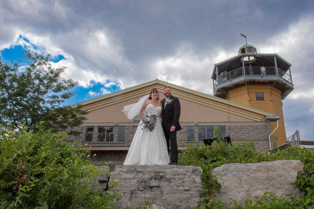 A bride and groom smile in front of a yellow tower and stone building on a cloudy day, dressed in formal wedding attire.