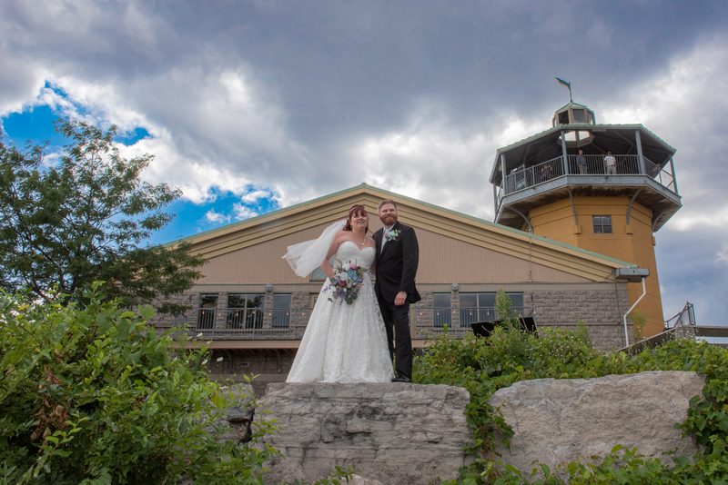 A bride and groom smile in front of a yellow tower and stone building on a cloudy day, dressed in formal wedding attire.