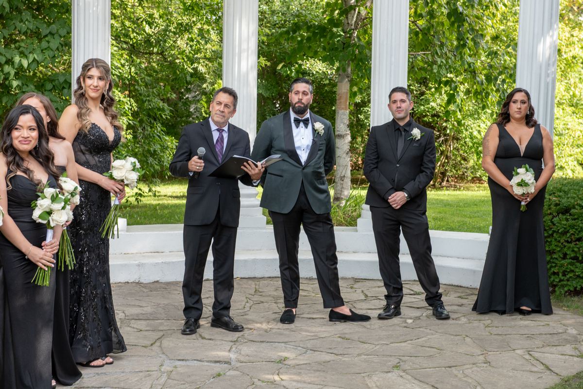 A wedding ceremony takes place outdoors under a white colonnade, with the officiant, groom, and bridesmaids in black gowns.