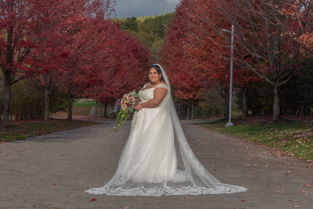 A bride in a white lace gown and veil stands on a path surrounded by crimson autumn trees, highlighting the rich fall colors.