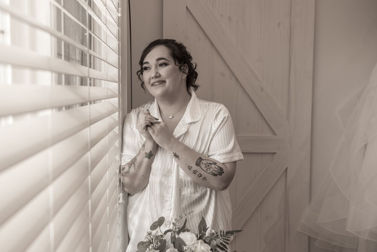 A bride in a white button-down and floral arrangement stands by a window, smiling gently as light filters through the blinds.