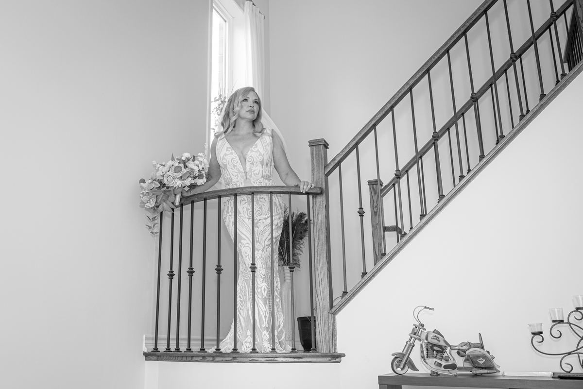 A bride in a white gown leans against a railing, gazing upward thoughtfully, illuminated by soft natural light from a window.