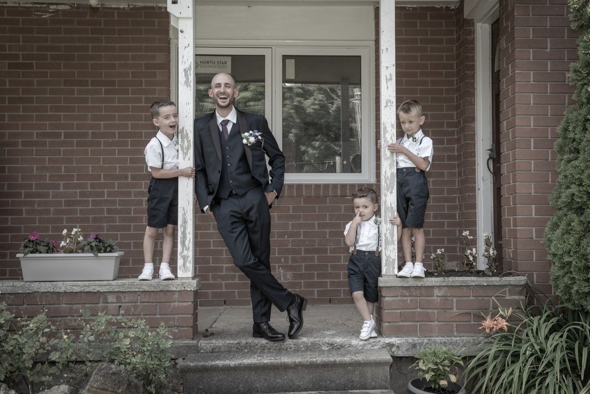 A laughing groom in a suit is flanked by three young boys on the steps of a brick building, bathed in soft daylight.