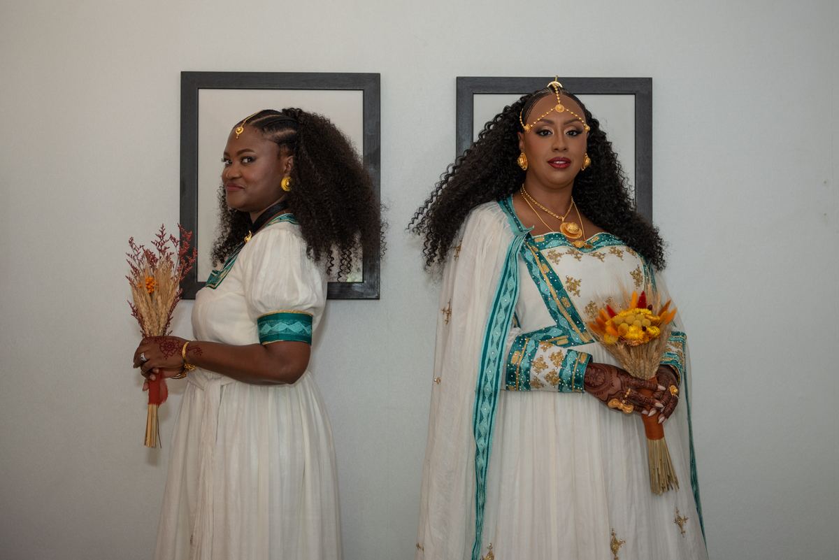 Two women in white and teal ceremonial dresses stand with flower bouquets and gold jewelry against a plain wall.