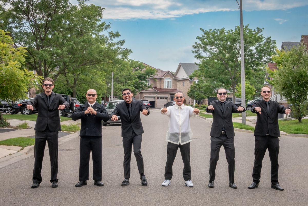 Six formally dressed men pose playfully on a suburban street, wearing sunglasses and making synchronized gestures.
