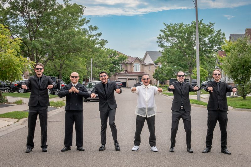 Six formally dressed men pose playfully on a suburban street, wearing sunglasses and making synchronized gestures.