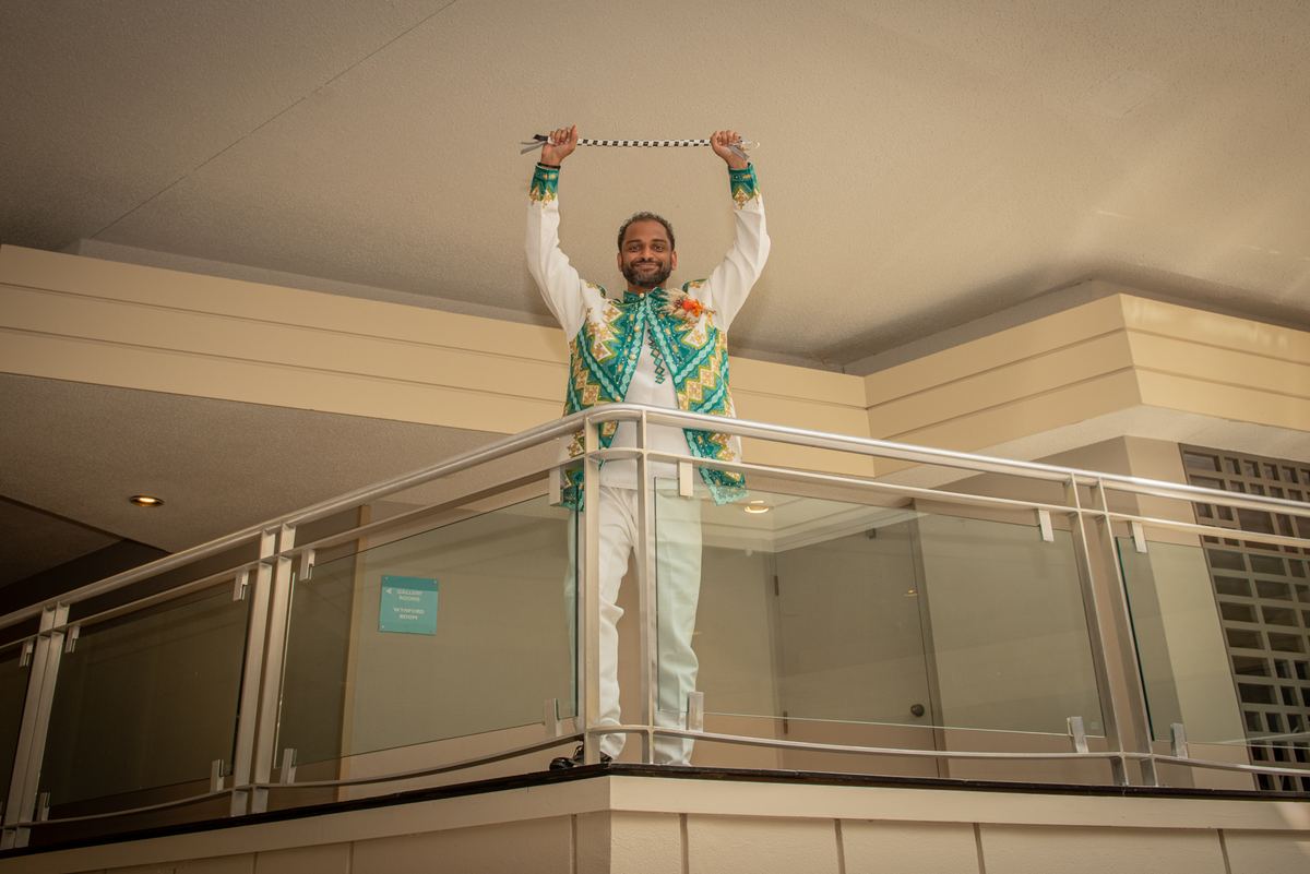 A man in an ornate white and green outfit stands on a balcony, arms raised with a ceremonial staff, exuding joy.