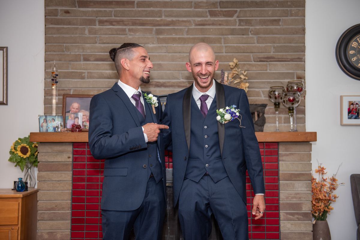 Two grooms in navy suits laugh by a brick fireplace with photos and decorations, creating a joyful indoor scene.