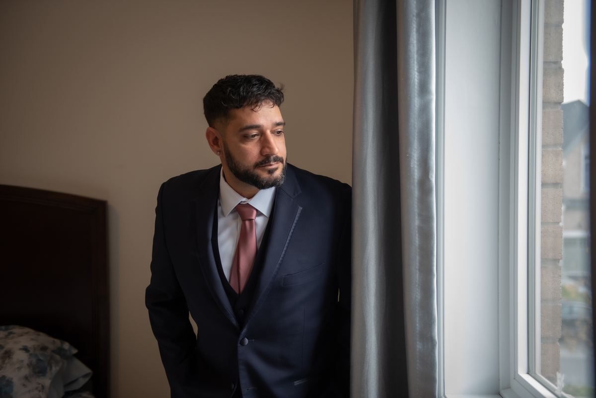 A man in a dark suit and pink tie stands by a window, gazing thoughtfully outside, illuminated by soft natural light.