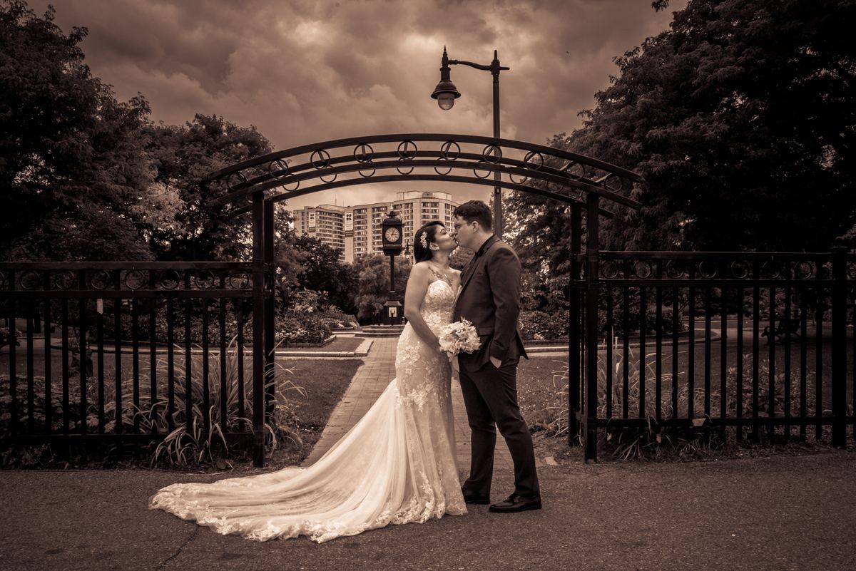 A newlywed couple kisses at a park gate under an ornate metal archway, framed by a moody, sepia-toned sky.
