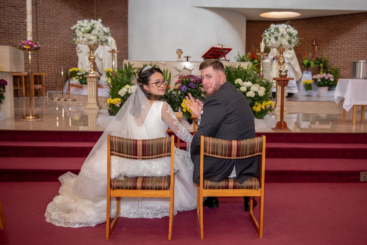 Bride and groom sit in wooden chairs, smiling and gesturing playfully, surrounded by soft light and floral arrangements.