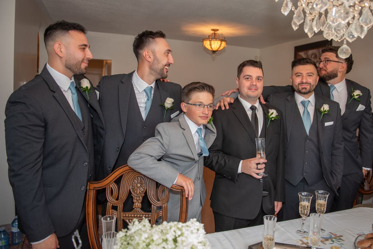 Groomsmen and a young boy in formal wear gather around a dining table, smiling and laughing with a champagne toast.