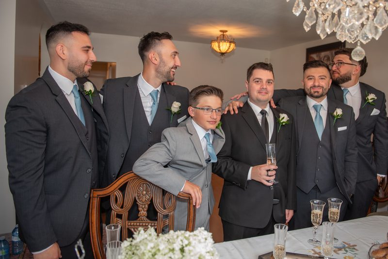 Groomsmen and a young boy in formal wear gather around a dining table, smiling and laughing with a champagne toast.