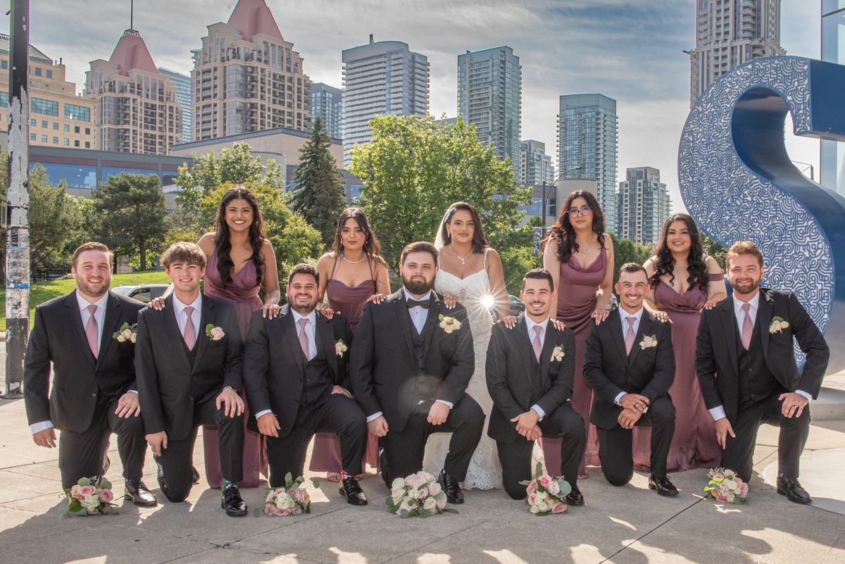 A wedding party poses outdoors with the bride and groom in front, surrounded by family members in black suits.