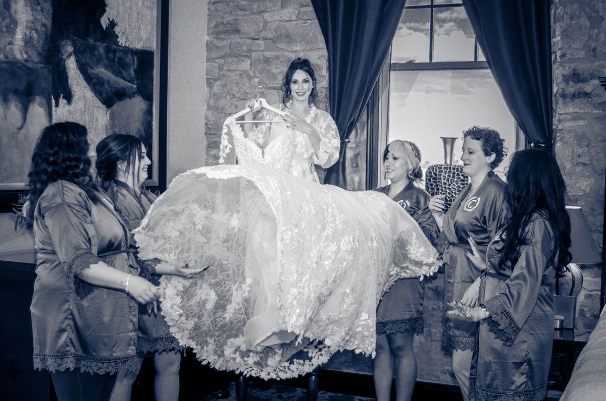 A bride smiles as her bridesmaids hold up her lace wedding gown in a joyful moment, lit by natural light.