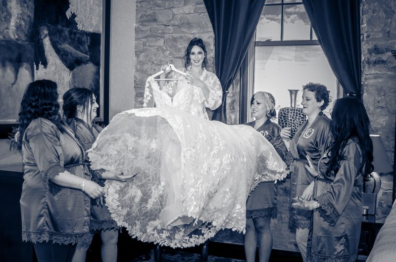 A bride smiles as her bridesmaids hold up her lace wedding gown in a joyful moment, lit by natural light.