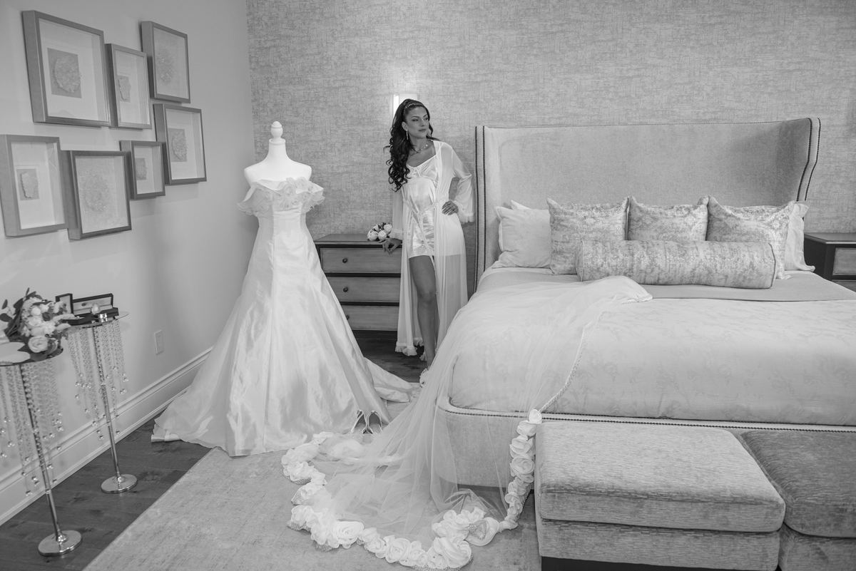 Bride stands beside mannequin in wedding gown, gazing thoughtfully at camera as dress train flows on floor.