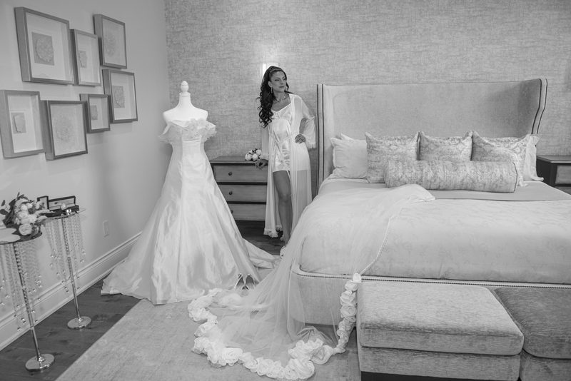 Bride stands beside mannequin in wedding gown, gazing thoughtfully at camera as dress train flows on floor.