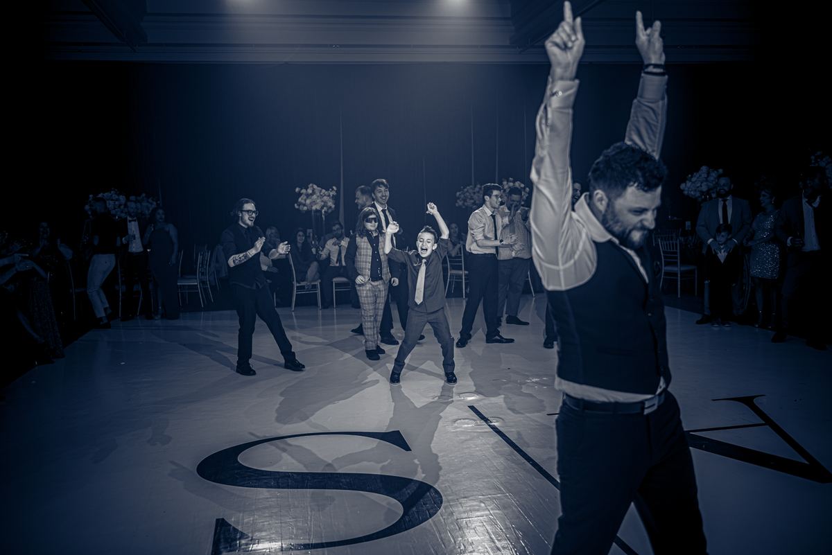 Guests, including young boys in suits, dance energetically on a decorated wedding reception floor under cool-toned lighting.