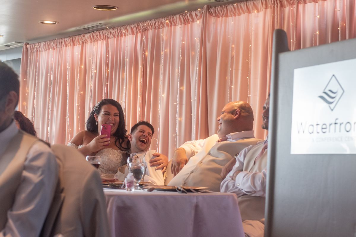 Bride and groom laugh as she records the moment at a warmly lit reception with guests in background.