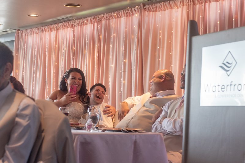 Bride and groom laugh as she records the moment at a warmly lit reception with guests in background.