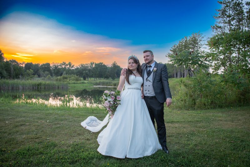 A bride and groom stand by a pond at sunset, their wedding attire reflecting the sky's orange and blue hues in the water.